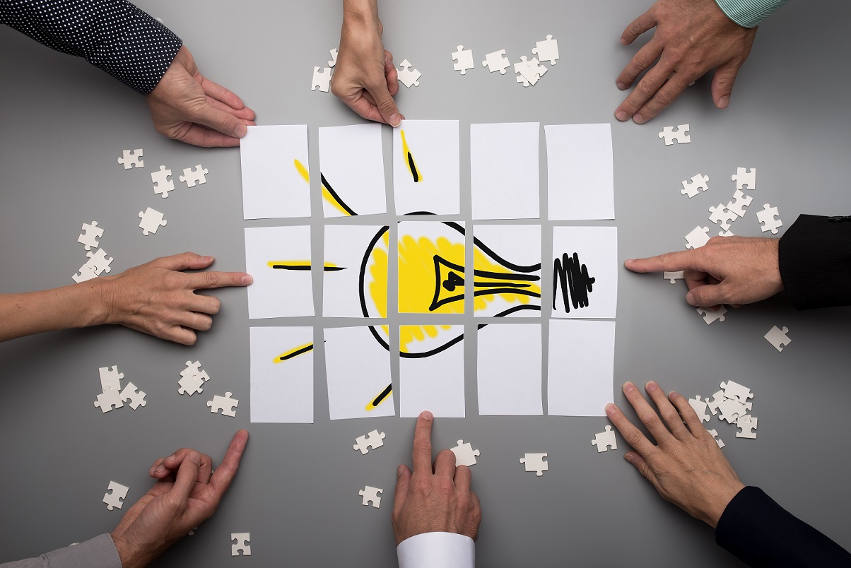 Top view of businesspeople hands touching white papers arranged on a gray table forming a yellow light bulb. Conceptual for brainstorming and teamwork.