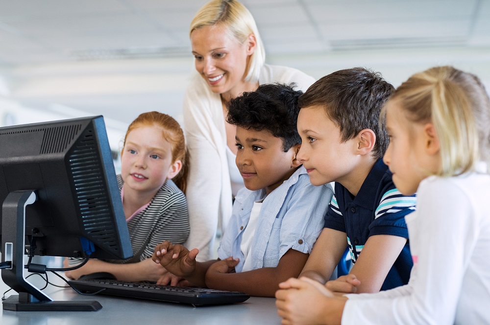 Teacher helping multiethnic children at computer terminal in primary school. Smiling school children looking at computer screen with teacher in library. Happy mature teacher and elementary students using desktop pc in classroom.